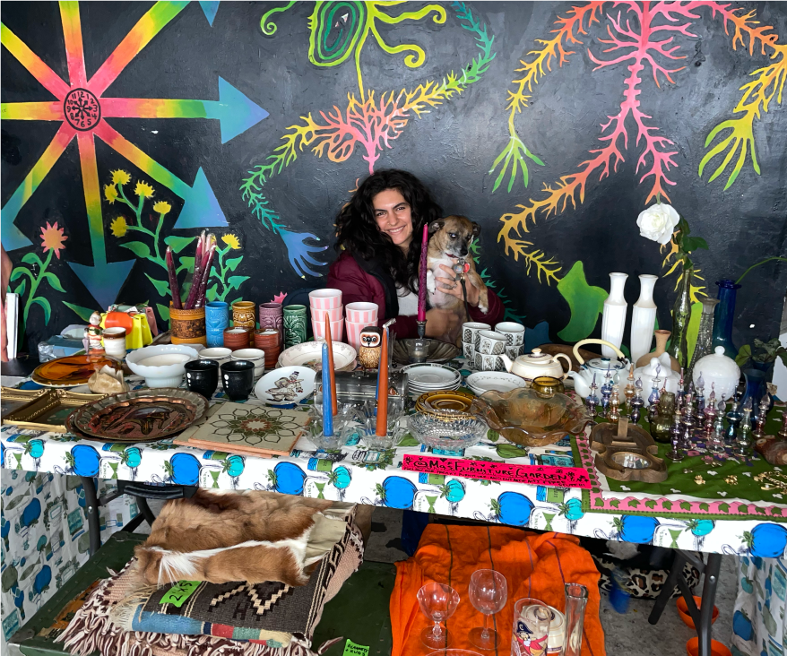Mantra Marie Bedross holding her dog while seated behind a big table full of old bowls and plates.