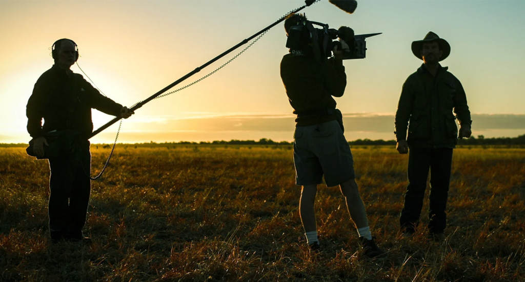 A film shoot on location with the outlines of a boom mic operator, a camera man, and an actor on an open plain to help illustrate what skills do you need to become a cinematographer.